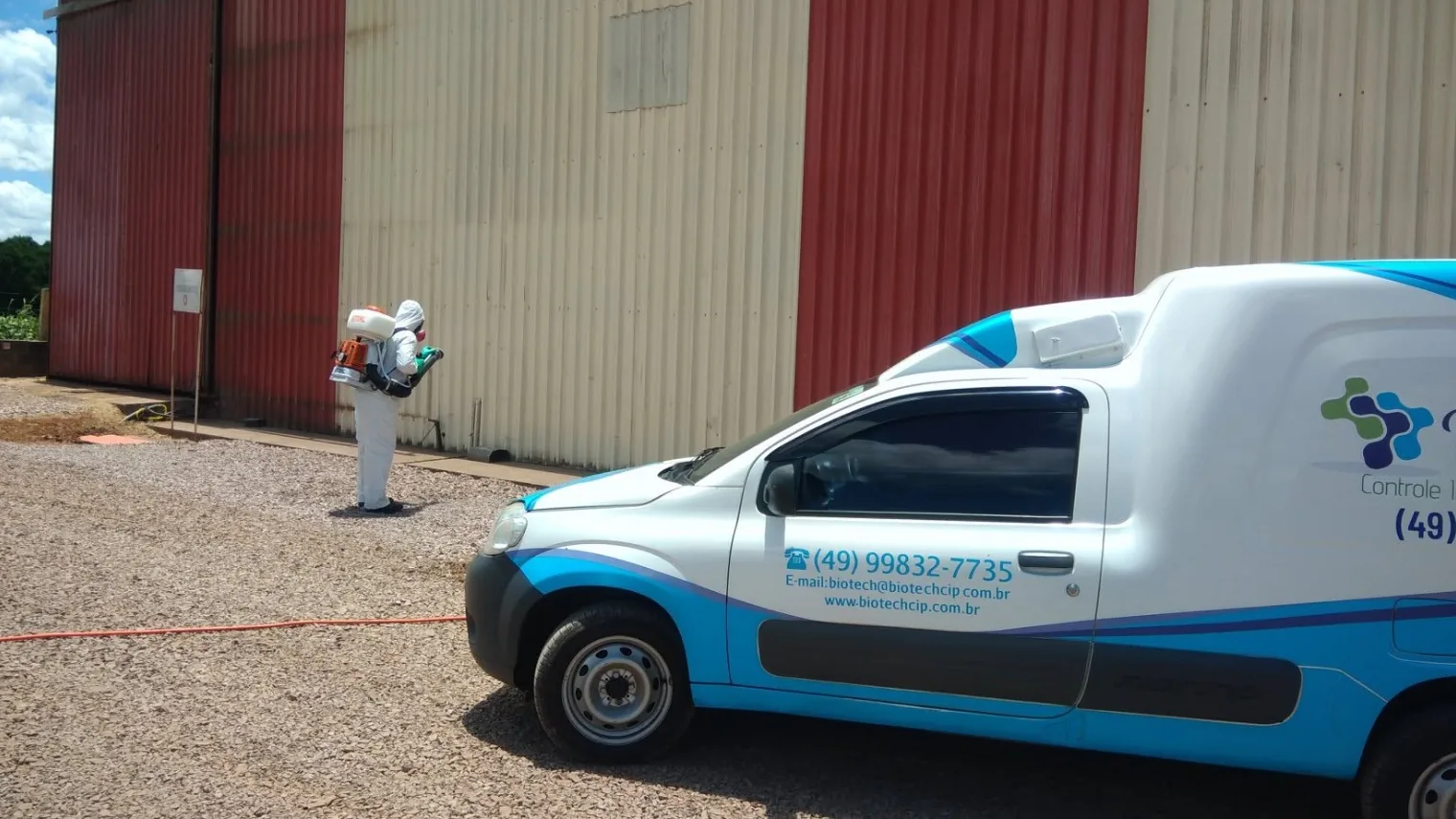 Veículo da Biotech em primeiro plano e, ao fundo, técnico de macacão branco integral aplicando com atomizador costal na parede de um barracão industrial.